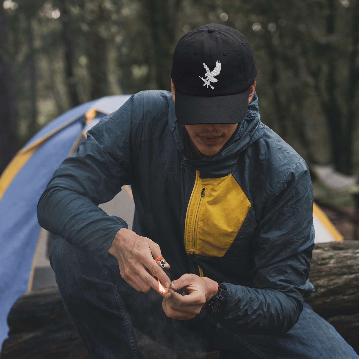 Person camping, sitting by a fire in a forest with a tent in the background, wearing black Rifle Eagle hat.