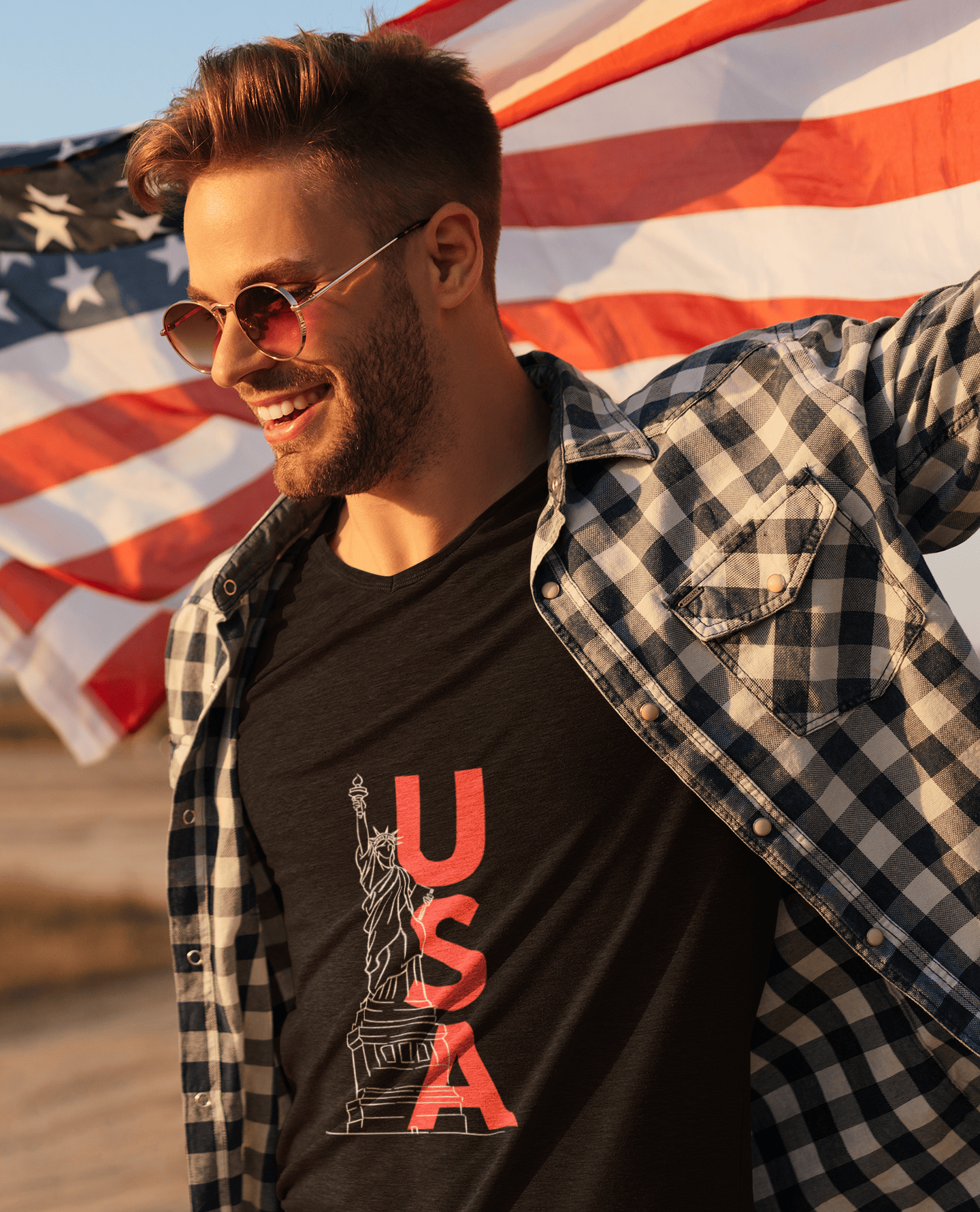 Man wearing sunglasses and a plaid shirt over a black t-shirt with 'USA' design, standing in front of an American flag.